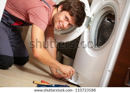 Young attractive smiling worker in uniform fixing washing machine, background