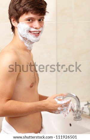 Young handsome attractive guy shave his beard while looking to the mirror, background