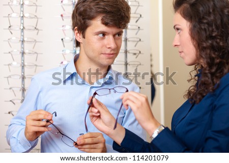 Attractive saleswoman helps young handsome guy buy eyeglasses, background in optician shop