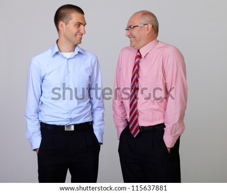 Happy smiling senior and junior businessman discuss something during their meeting with hands in pockets, isolated on grey