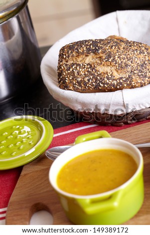Bowl of soup on kitchen top placed on red mat with steel pot and basket full of bread