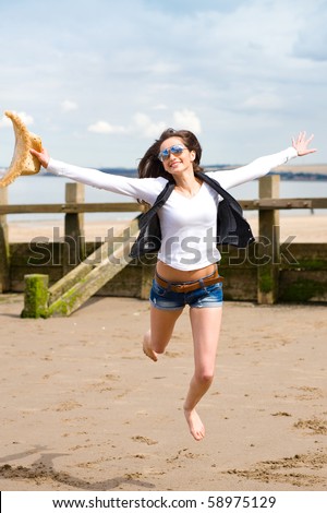 young attractive female jump high over sandy summer, wears sunglasses and holds straw hat in her hand