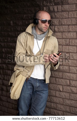 young male in khaki jacket listen to his music, lean against wall, outdoor shoot