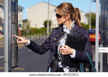 Attractive and trendy businesswoman wait for bus, check bus time on bus stop timetable, street shoot