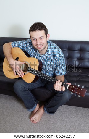 Young attractive male sits on sofa and plays acoustic guitar, indoor shoot