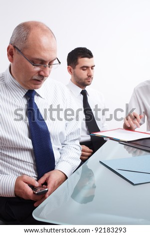 Two businessman, one mature texting using his mobile phone and one young sitting at table during meeting