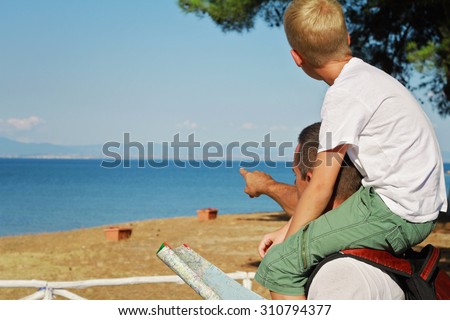 Father giving his son piggyback ride outdoors. Man and boy son looking at map in front of sea, pointing away, active summer holiday vacation, family travel photo