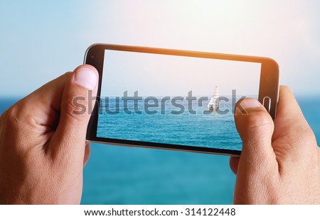 Male hand taking photo of Sailboat, sea, sun, blue, sea, horizon and ocean with cell, mobile phone.