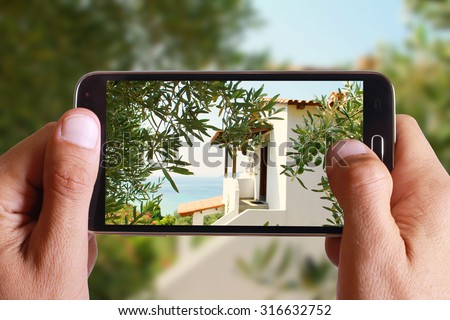 Male hand taking photo of Idyllic Greek house, villa, by the sea , between olive trees with cell, mobile phone. Greece holiday.