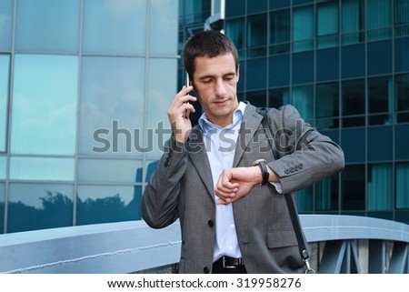 Young handsome, successful businessman talking on the phone in the city, in front of modern building. Businessman in a hurry looking at his watch