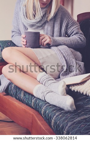 Beautiful Woman wearing  cozy sweater and Warm Wool Socks  reading a book sitting on comfortable sofa enjoying hot chocolate drink . Winter, Christmas  holidays concept.