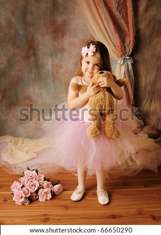 Adorable little girl dressed as a ballerina in a tutu, hugging a teddy bear standing next to pink roses.