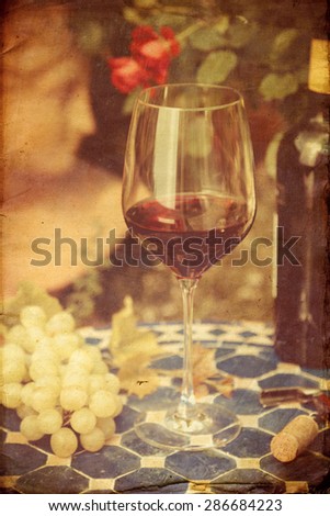 vintage style picture of a still life of a glass red wine with wine grapes on a table with a romantic garden in the background