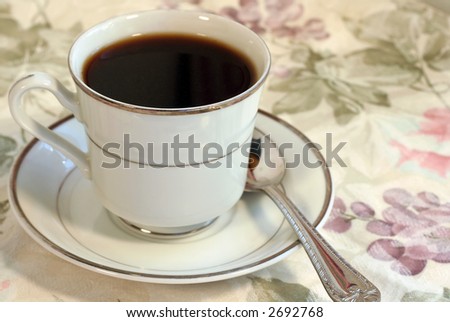 A cup of coffee on a saucer with a floral table cloth.