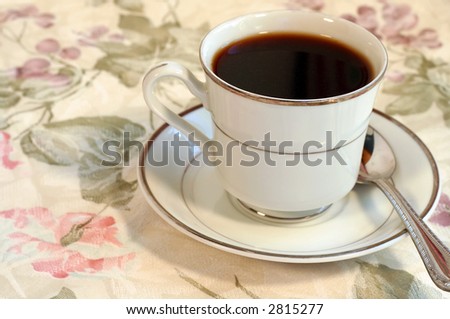 A cup of coffee on a saucer with a floral table cloth.