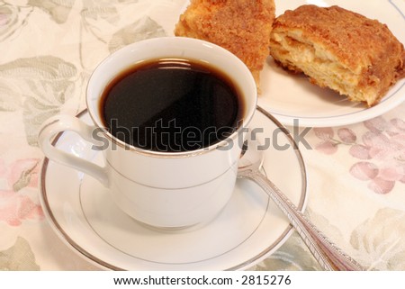 A cup of coffee on a saucer with a floral table cloth with sugar topped sweet rolls.