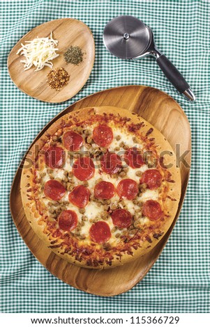 Pizza on a wooden round pizza stone with pizza cutter and grated cheese on the side