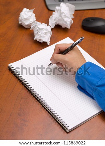 Close-up image of human hand writing on spiral writing pad on wooden office desk with crumpled papers.