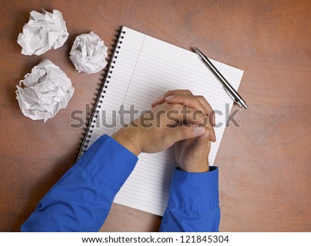 Close-up shot of clasped human hand with spiral notepad and pen with crumbled paper on wooden desk.