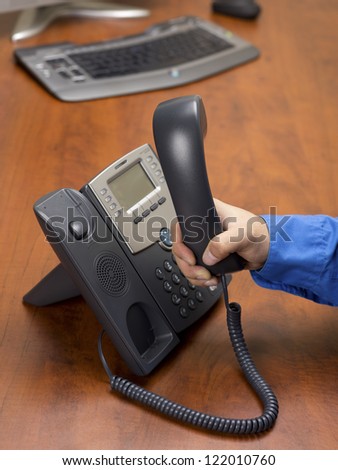 Close-up shot of a person holding black land line phone receiver in hand on wooden desk.