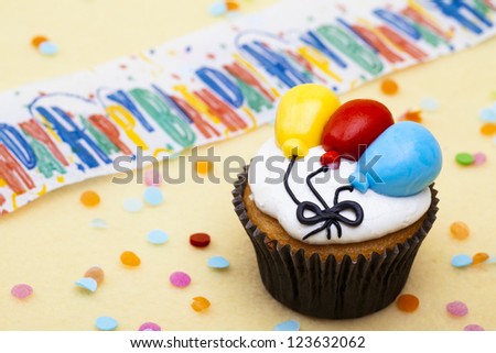 Close-up shot of a cupcake with colorful balloon shape and happy birthday sign in background.