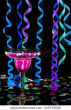 Close-up shot of martini with pink drink and streamers and colorful confetti over dark background.