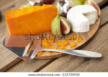 Close-up shot of cheese slicer with cheese and dig fruit on wooden board.