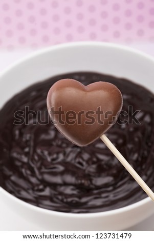 Closed up shot of a heart shape chocolate in a stick with blurred chocolate dip background