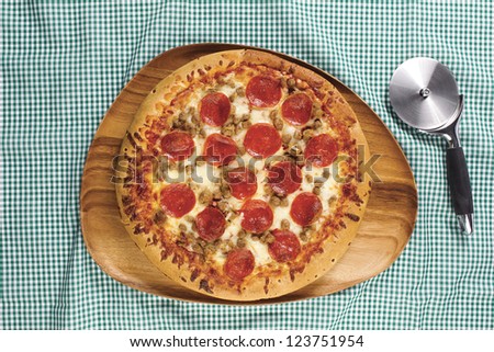 Close-up overhead shot of pepperoni pizza in wooden plate with pizza cutter on table cloth.