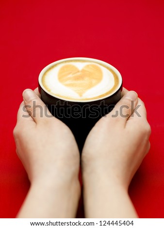 Two hands reaching for a coffee cup, with a heart on top of the cappuccino foam.