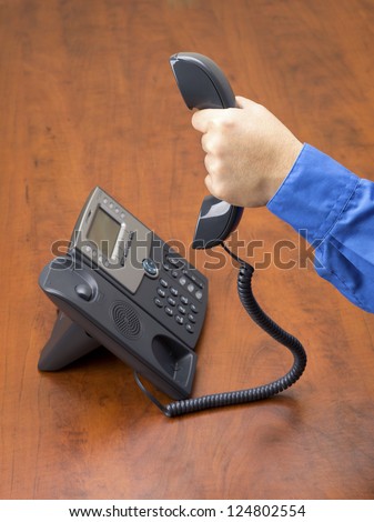 Detailed shot of a human hand holding land line phone receiver in hand on wooden office desk.