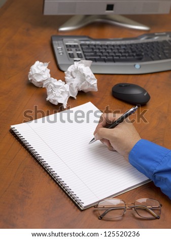 Close-up shot of human hand writing on spiral notepad on wooden office desk with spectacles and crumpled papers.