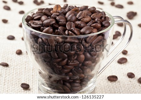 Pile of coffee beans in a transparent pitcher with scattered pieces on the table cloth