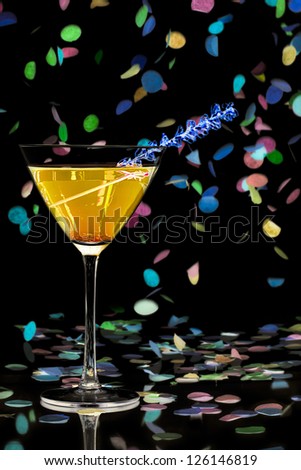 Close-up shot of martini glass with yellow cocktail and colorful decorative confetti against plain black background.