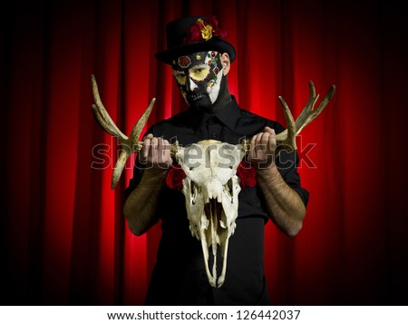 Portrait shot of a scary male with sugar skull paint posing with animal skeleton in hand over red background.
