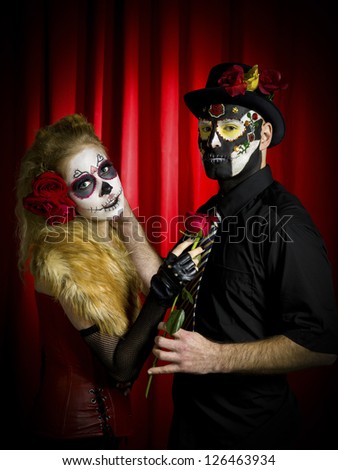 Portrait shot of ugly looking man with woman wearing sugar skull make up and holding rose over red background.