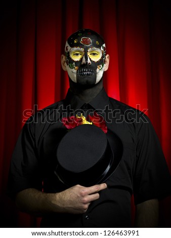 Portrait shot of a ugly man wearing sugar skull paint posing with hat in hand.