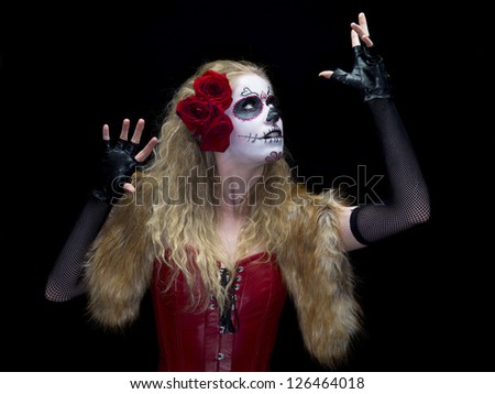 Image of a scary woman wearing sugar skull face make-up posing against dark background with hand raised.