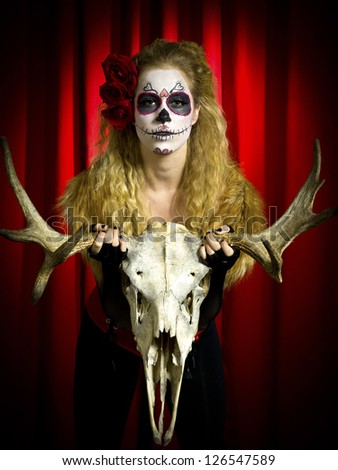 Portrait shot of a scary woman wearing sugar skull holding animal skull over red background.