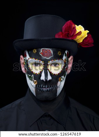 Portrait shot of a scary man wearing traditional sugar skull make-up with hat and roses against black background.