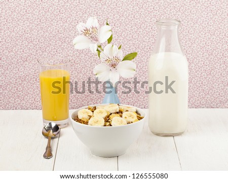 Bowl of cereals with banana toppings together with a spoon, glass of orange juice, bottle of fresh milk and base with flowers