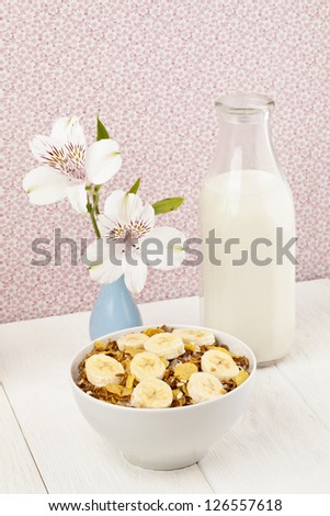 Close up image of bowl of cereal with banana,  bottle of milk and flower on wooden table