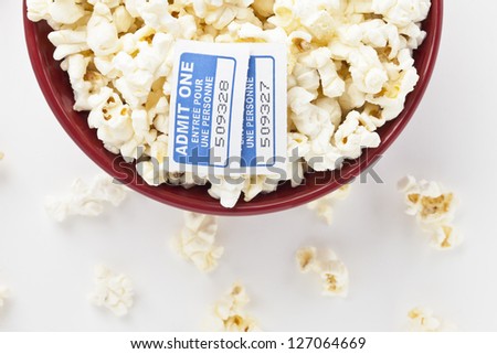 Cropped image of a red bowl with popcorn and two movie tickets laid in a white background with scattered popcorn pieces