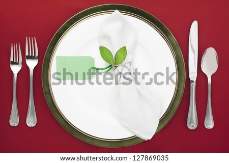 Silver utensils with plate and ring napkin with green card and leaf on the red table background