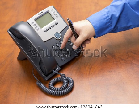 Close-up shot of human hand dialing number on black land line phone on wooden desk.