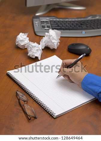 Close-up shot of a person writing on spiral notepad on wooden desk with spectacles and crushed papers.