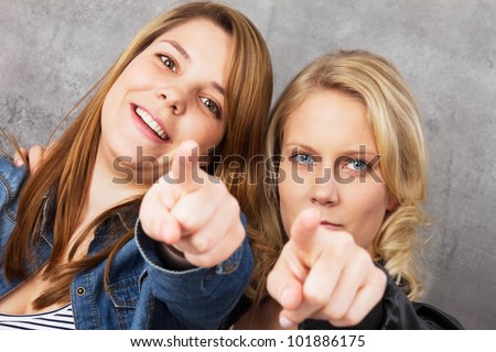 We want you - portrait of two young women pointing with their fingers at the camera. Studio shot against a gray background.