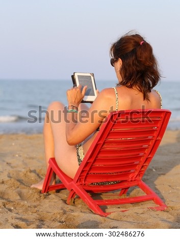 tanned woman reads the ebook on the seashore in the summer while his children playing on the beach