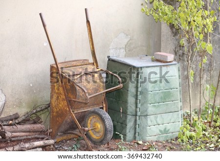 old wheelbarrow in the garden and the big green container for compost to be used as natural fertilizer for the garden ecology