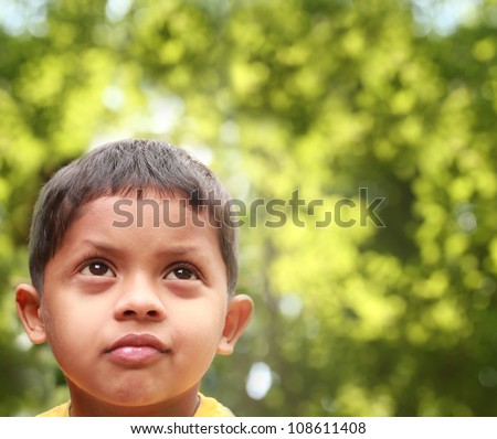 Young indian boy of kinder-garten school age thinking or dreaming about playing and having fun after being bored. Background is blurred trees in the backdrop acting as copy-space.